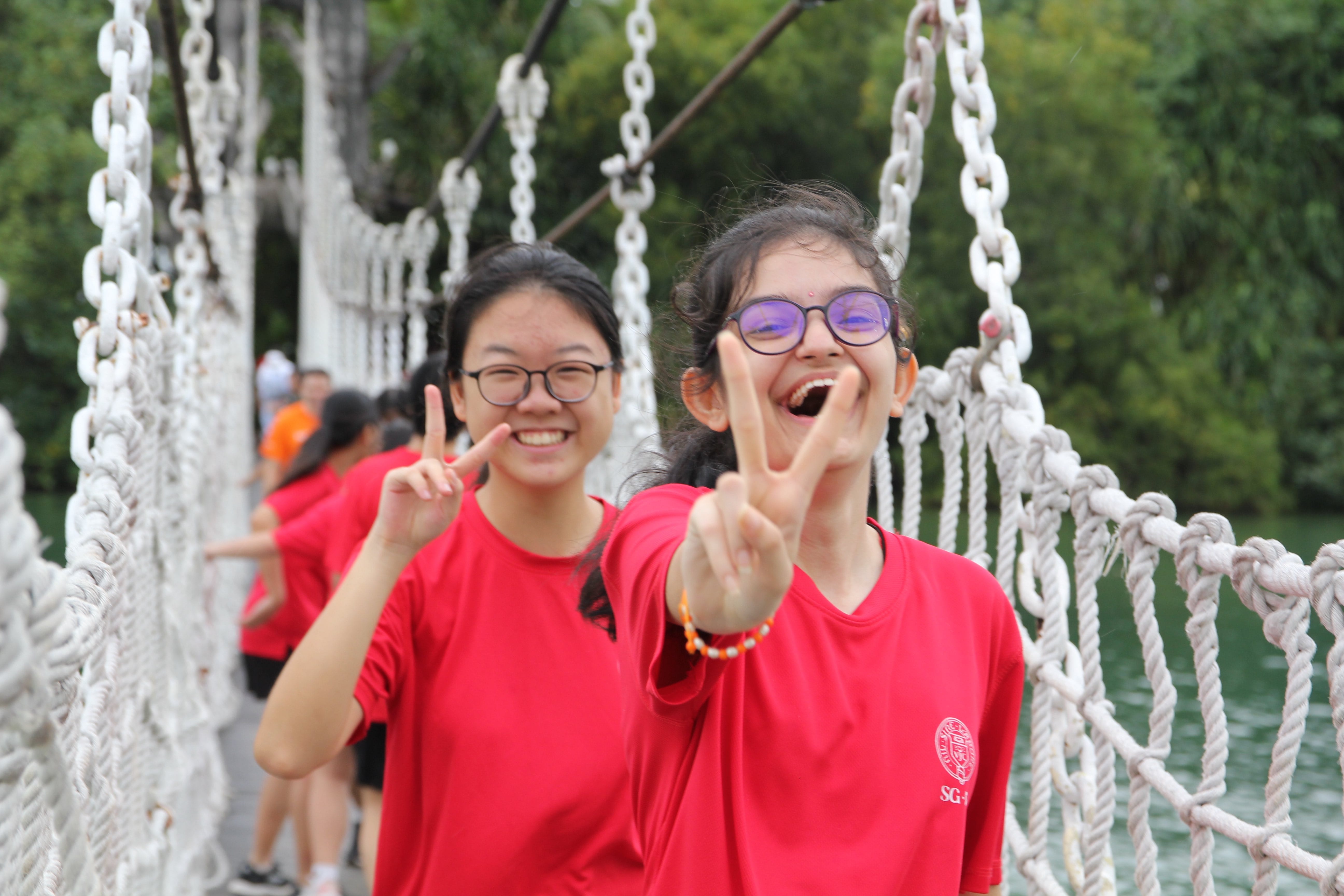 Two smiling girls in red shirts make peace signs on a rope suspension bridge surrounded by green trees.